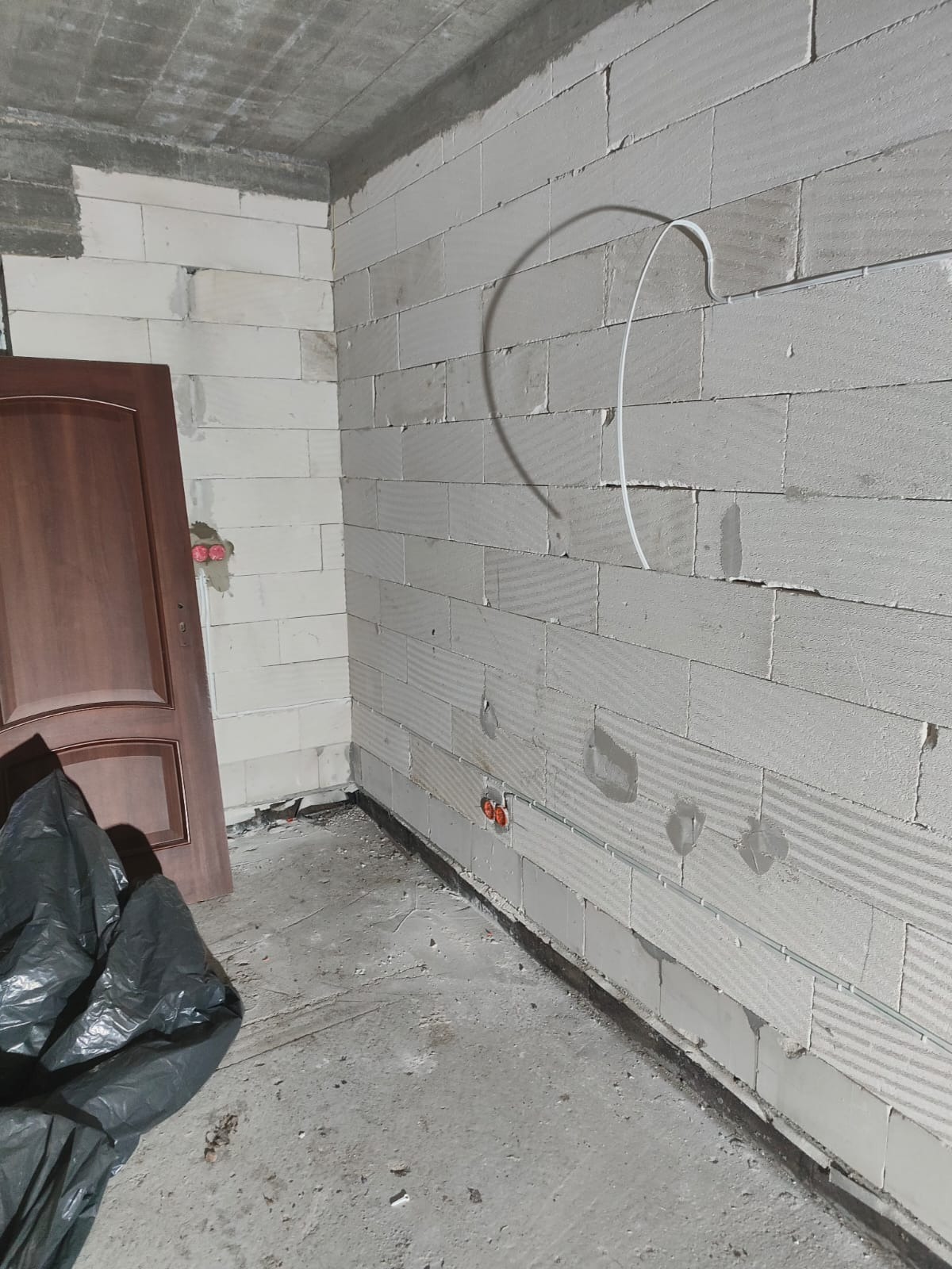 Wnętrze w trakcie remontu: ściana z białych bloczków, częściowo położona instalacja elektryczna w peszlu, widoczne puszki instalacyjne, surowy betonowy sufit, drzwi z ościeżnicą oparte o ścianę...