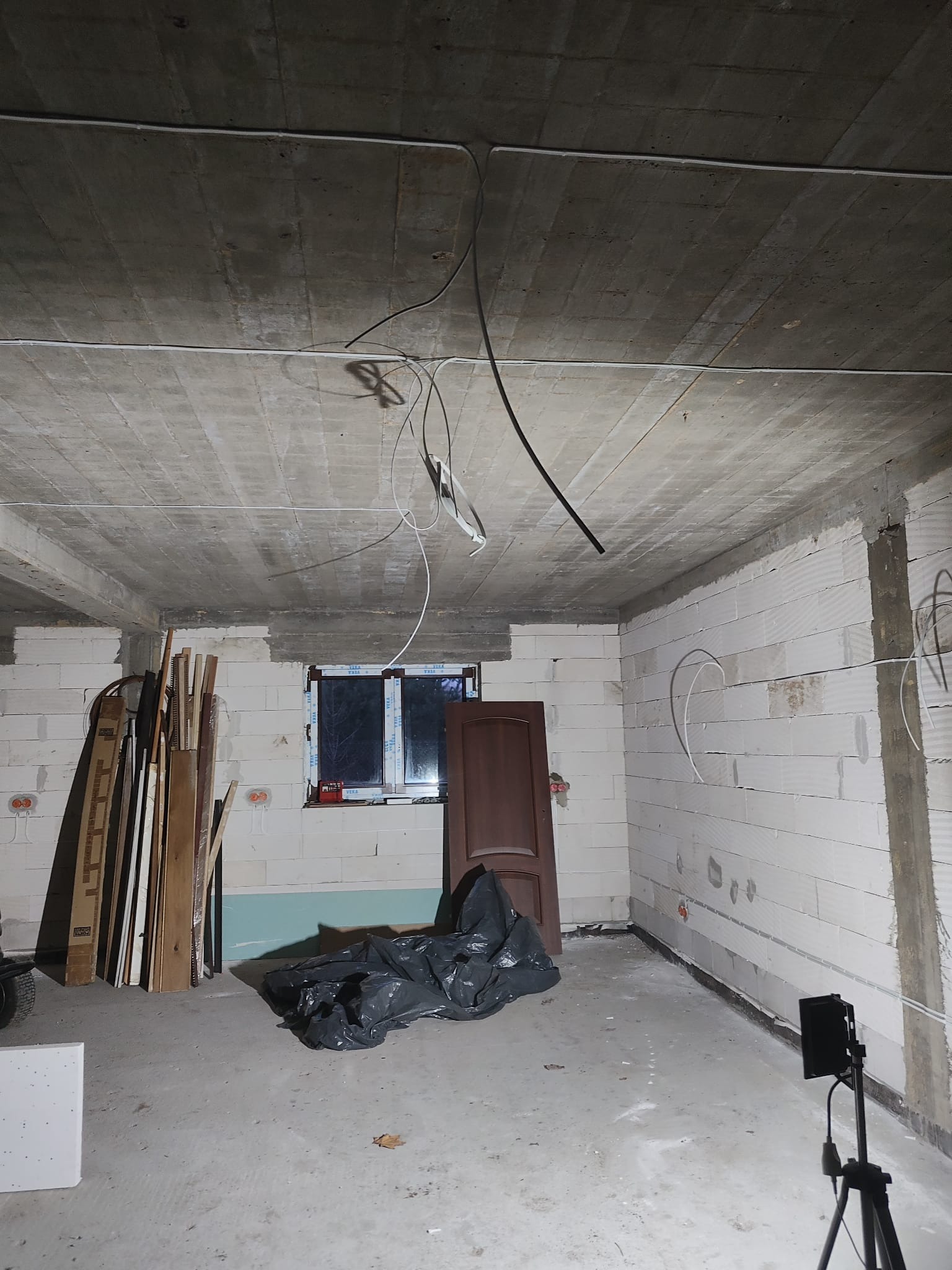 Surowy stan piwnicy w trakcie remontu, z widocznymi przewodami elektrycznymi wystającymi z sufitu i ścian, pustaki na ścianach, okno z parapetem, drzwi wewnętrzne oparte o ścianę, oraz materiały...