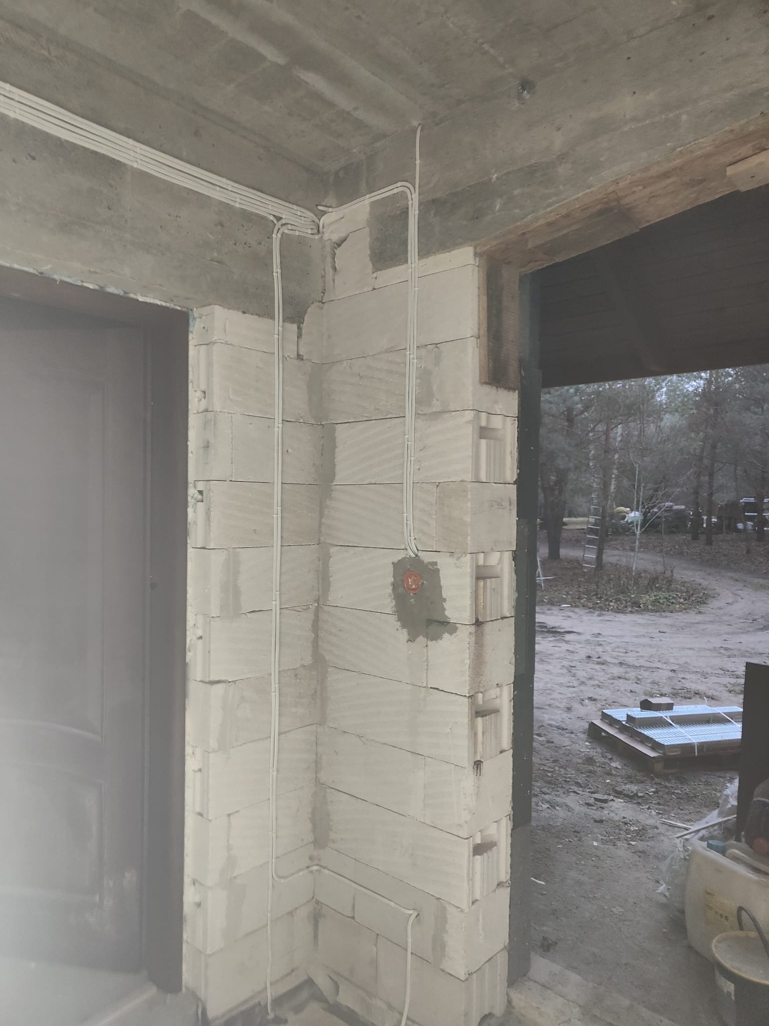 Szare przewody elektryczne poprowadzone po ścianie z betonu komórkowego w surowym wnętrzu budynku w trakcie budowy. Widoczna puszka elektryczna w narożniku i fragment drzwi.