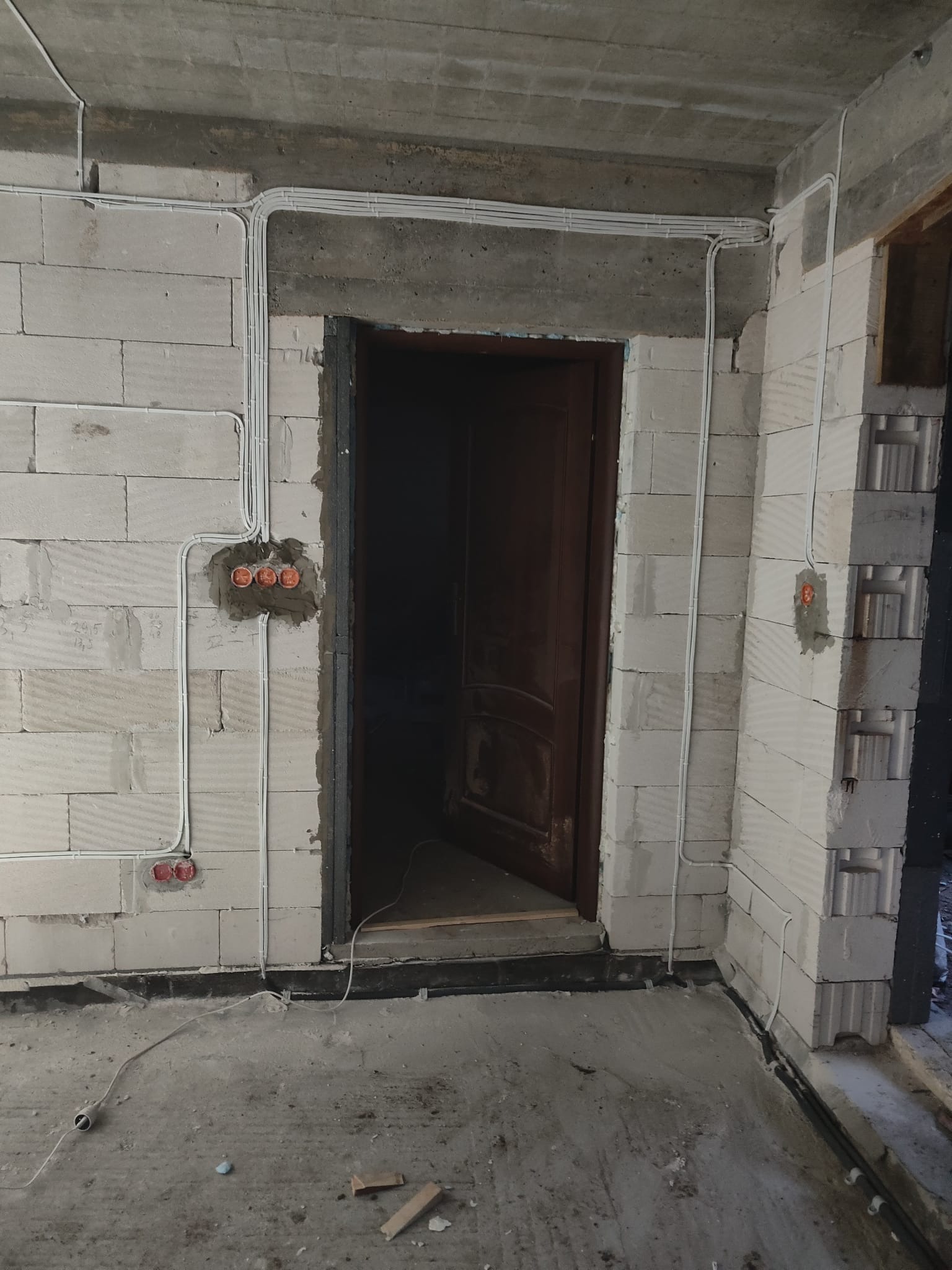 Wnętrze w trakcie remontu z widoczną instalacją elektryczną: białe przewody ułożone na ścianach z betonu komórkowego, puszki elektryczne w ścianie, surowy betonowy sufit, otwór drzwiowy z ciemnymi...