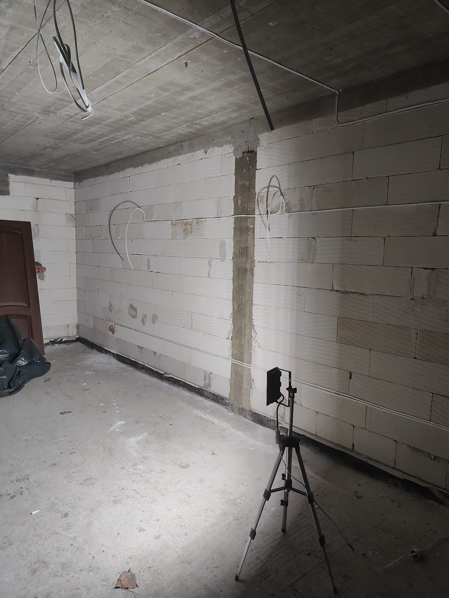 Surowy stan wnętrza z białymi ścianami z betonu komórkowego, widoczne kable elektryczne prowadzone po ścianach i suficie, oraz lampa na trójnogu oświetlająca przestrzeń.