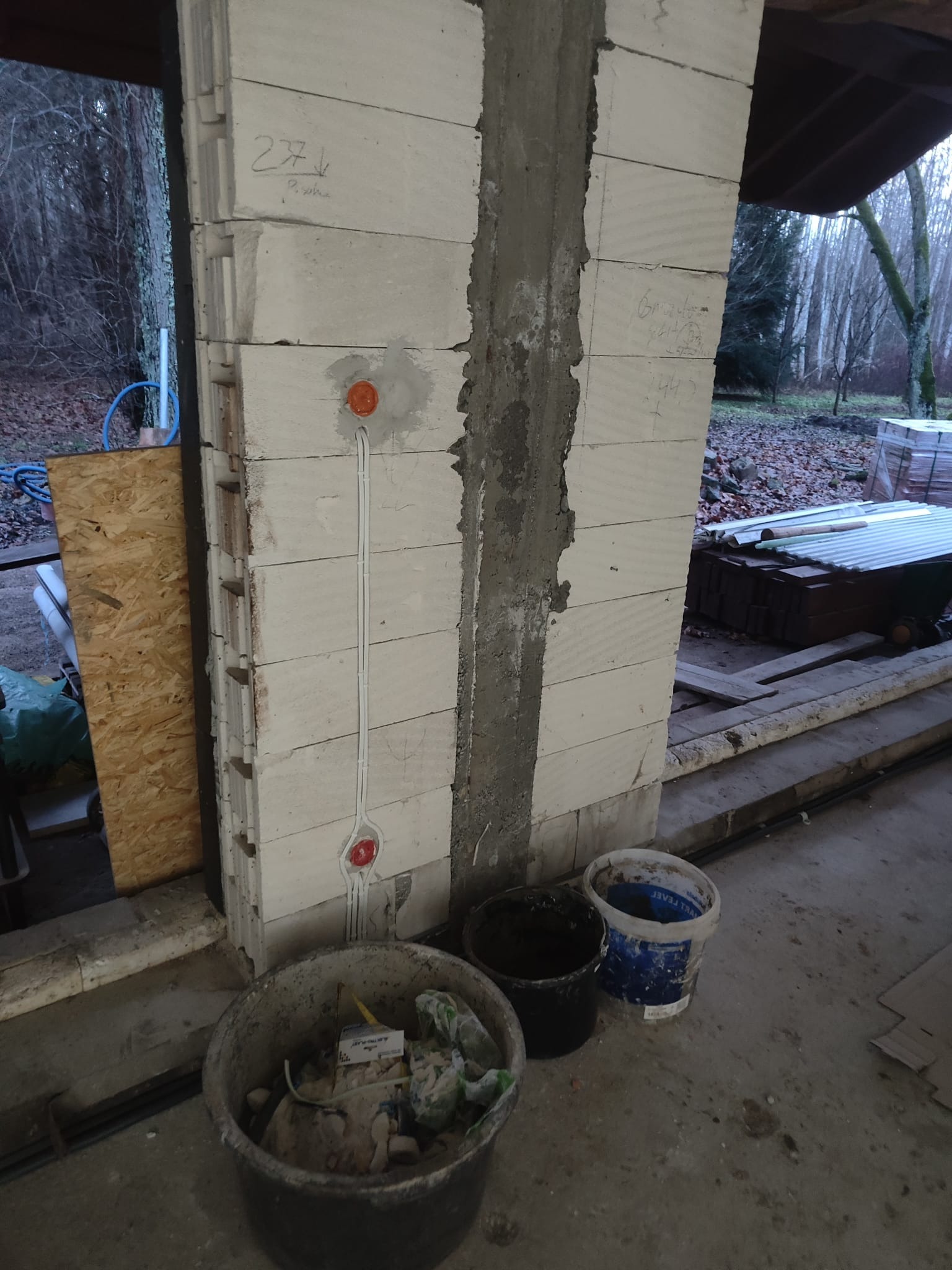 Wewnętrzna ściana z betonu komórkowego w trakcie budowy, z wyfrezowanymi kanałami na przewody elektryczne i zamontowanymi puszkami instalacyjnymi, widoczne wiadra budowlane i materiały.