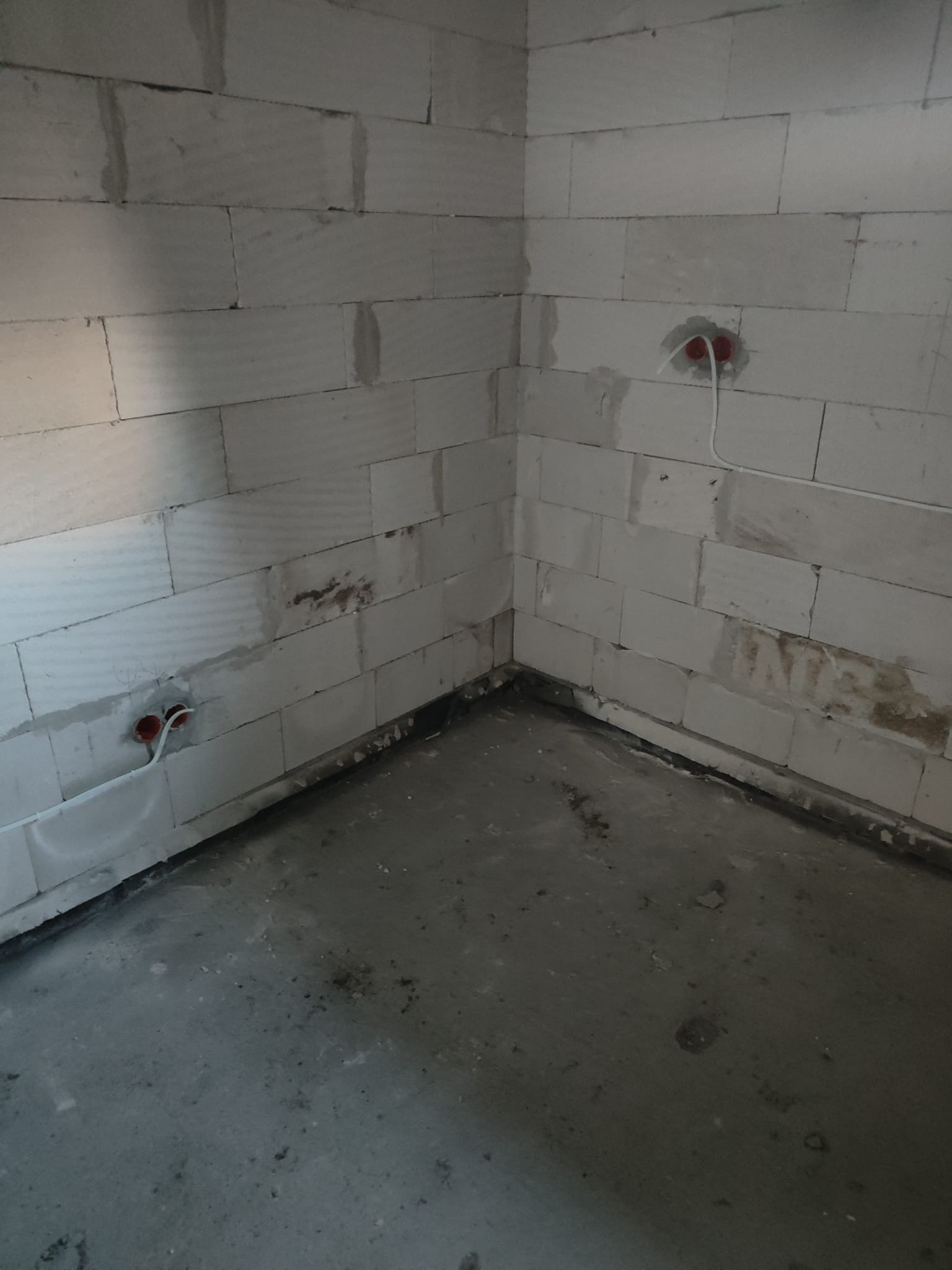 Surowy kąt pomieszczenia z bloków betonowych z dwoma puszkami elektrycznymi i wystającymi przewodami, widoczny szary betonowy podkład.