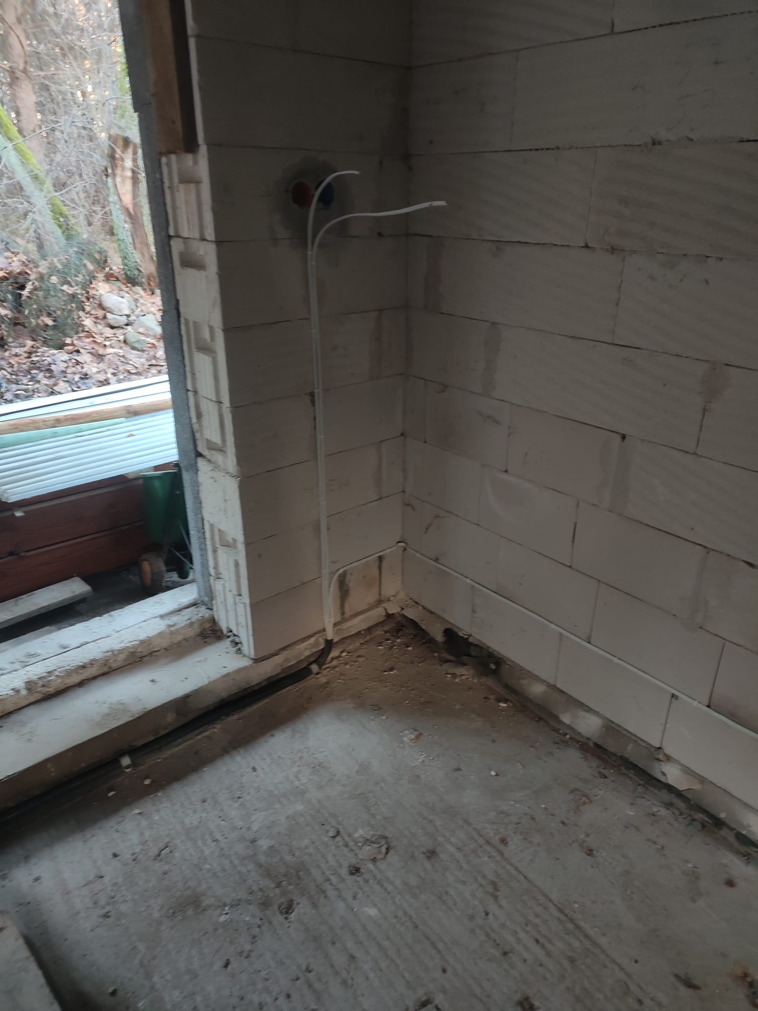 Surowe wnętrze w trakcie remontu: ściany z bloczków, wystające przewody elektryczne z puszek, surowy beton na podłodze, widok na zewnątrz przez okno, w tle drzewa bez liści.