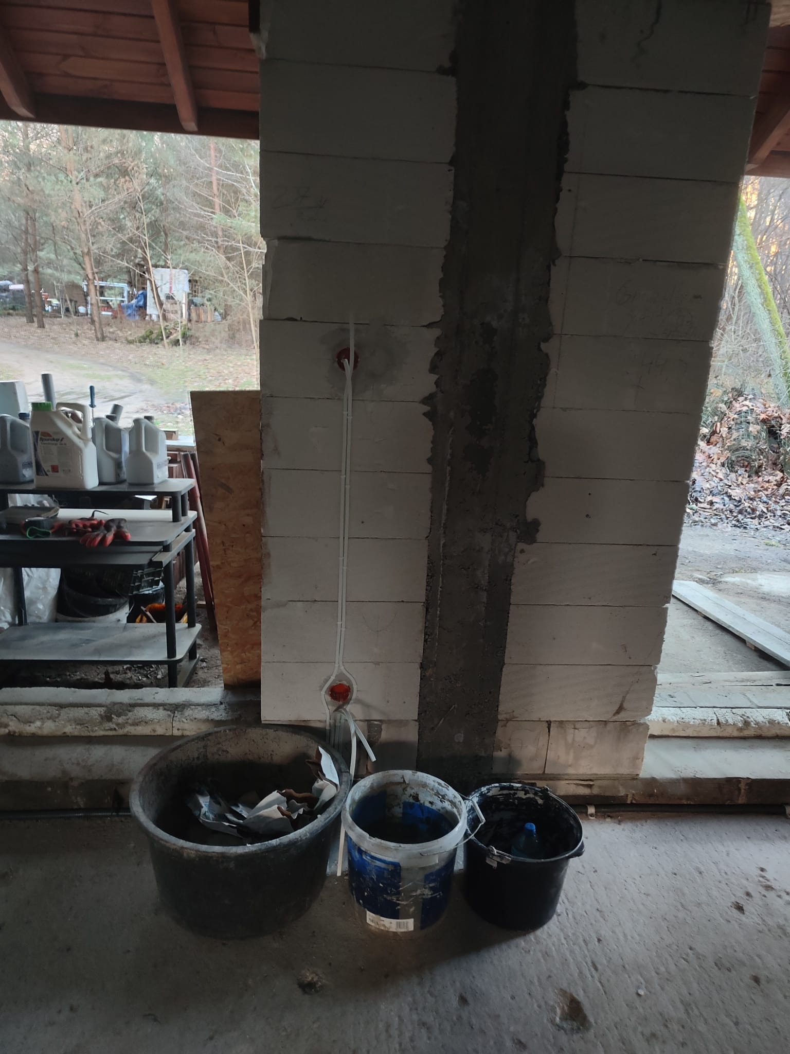 Wewnętrzna ściana z betonu komórkowego w trakcie budowy, z pionowym bruzdowaniem wypełnionym zaprawą, w którym poprowadzono instalację elektryczną z puszkami pod gniazdka. Na podłodze widoczne trzy...