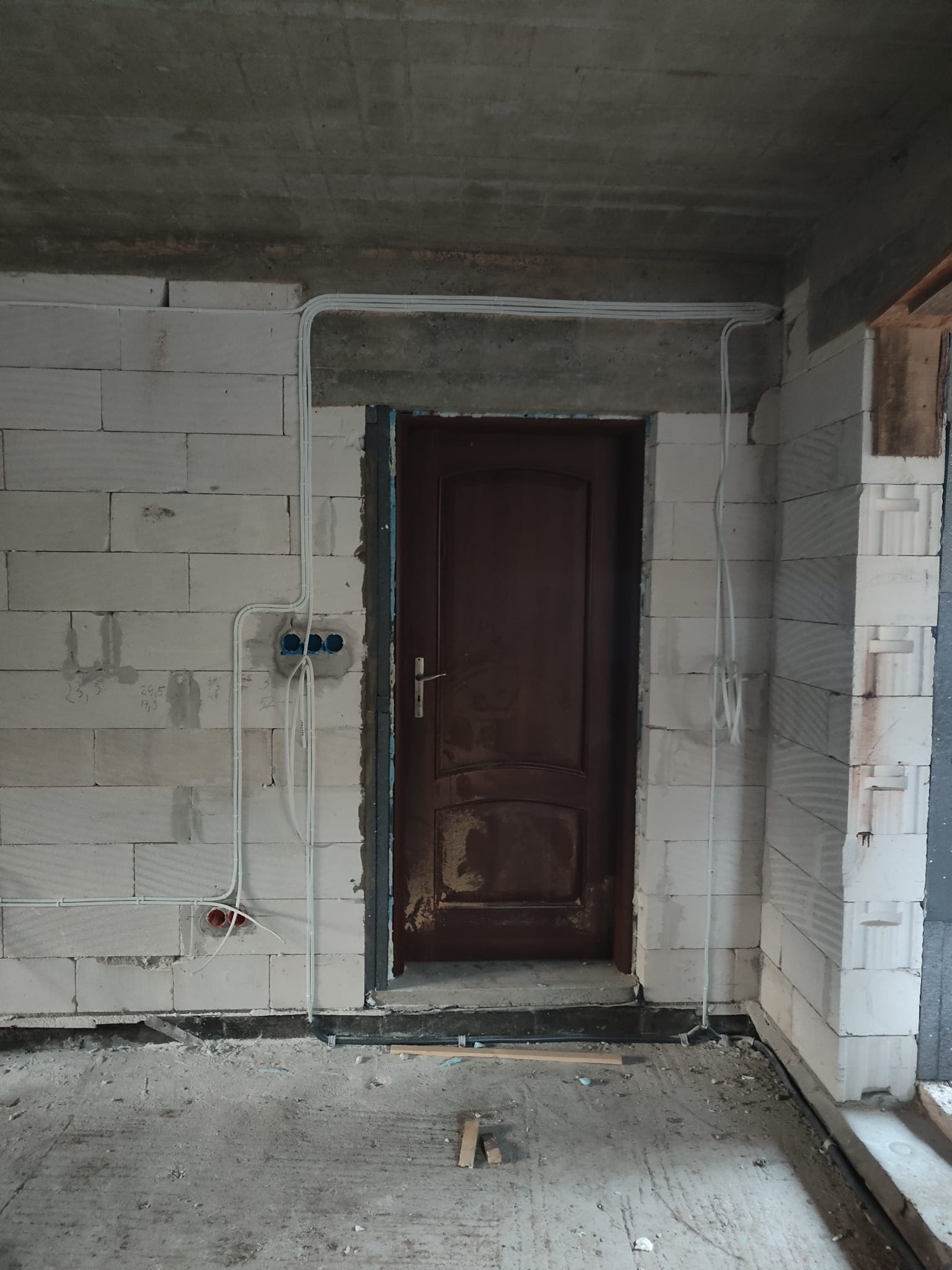 Surowe wnętrze w trakcie remontu z widocznymi przewodami elektrycznymi poprowadzonymi po ścianach z białych bloczków, puszki instalacyjne zamontowane w ścianie, stara, drewniana ościeżnica i drzwi.