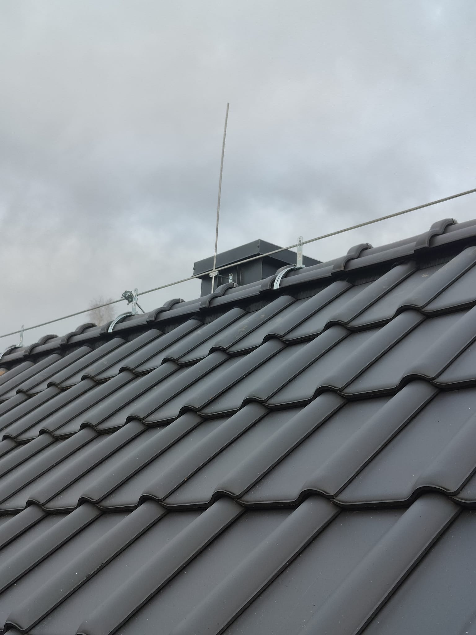 Szare dachówki ceramiczne z zamontowaną instalacją odgromową: przewód uziemiający z uchwytami i maszt odgromowy na tle pochmurnego nieba.