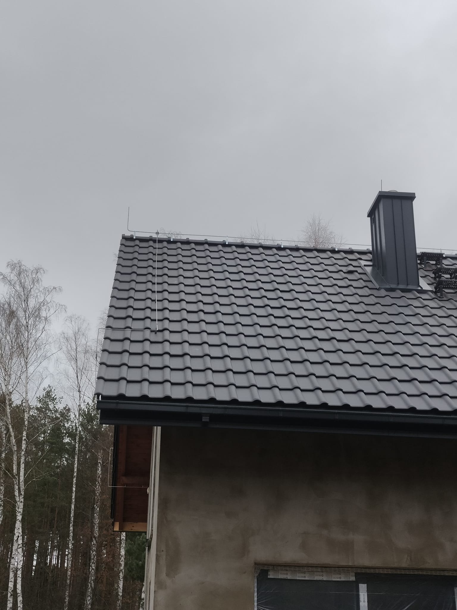 Szary dach z metalowej dachówki z zamontowaną instalacją odgromową, widoczny komin i fragment ściany budynku w trakcie wykończenia, tło stanowią drzewa i pochmurne niebo.