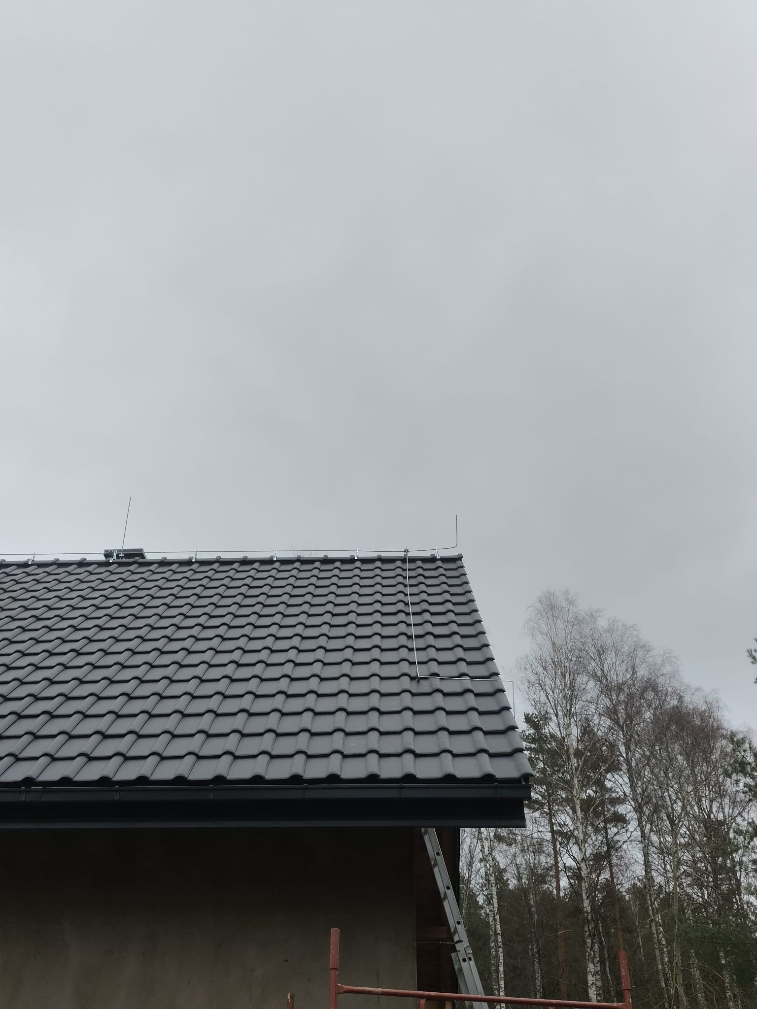 Szary dach z dachówki ceramicznej z zamontowaną instalacją odgromową, widoczne przewody i maszty, w tle drzewa i pochmurne niebo.