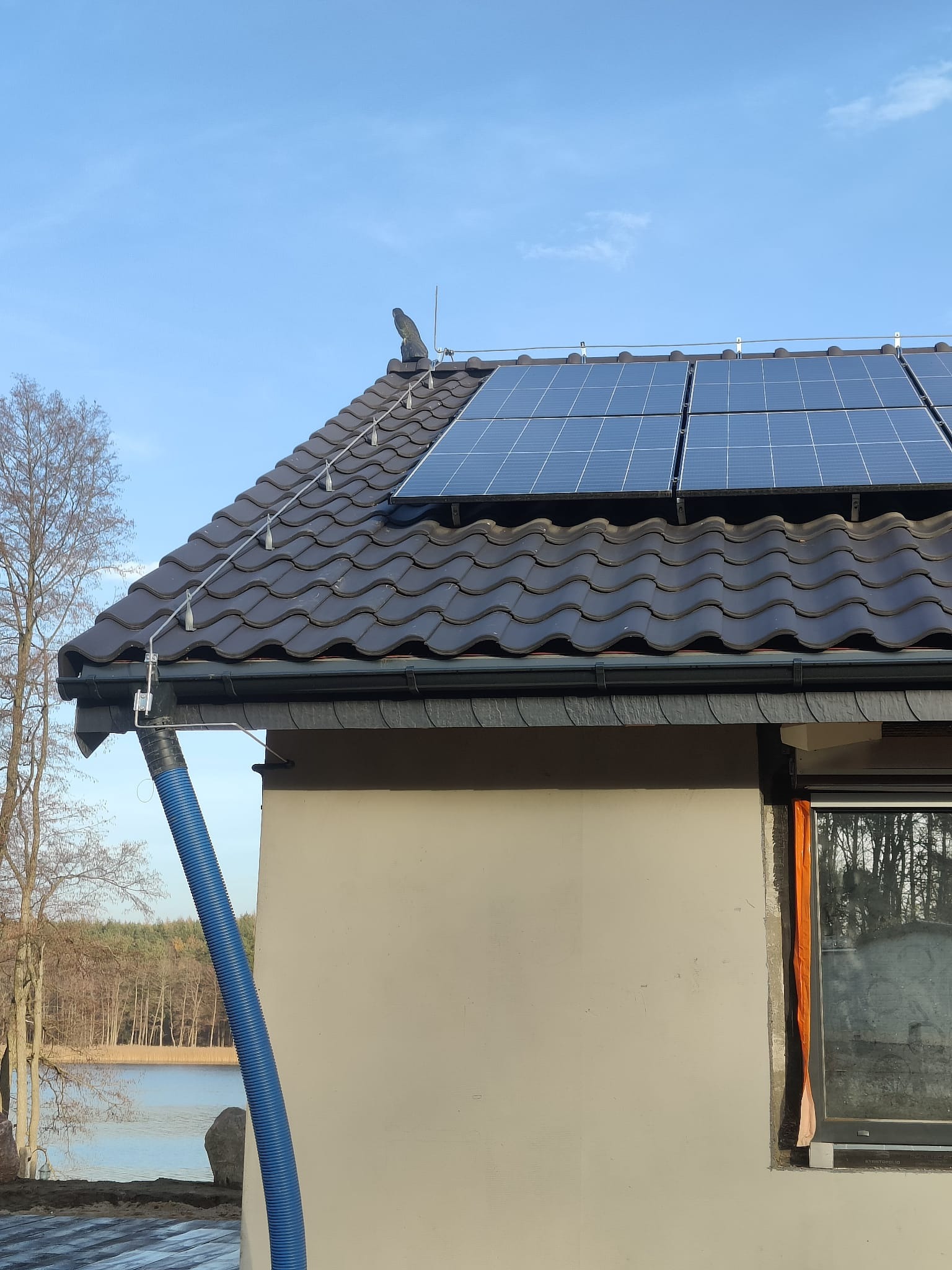 Brązowy dach z dachówki ceramicznej z zamontowanymi panelami fotowoltaicznymi i systemem odgromowym, niebieski przewód wentylacyjny na elewacji budynku, widok na jezioro i las w tle.