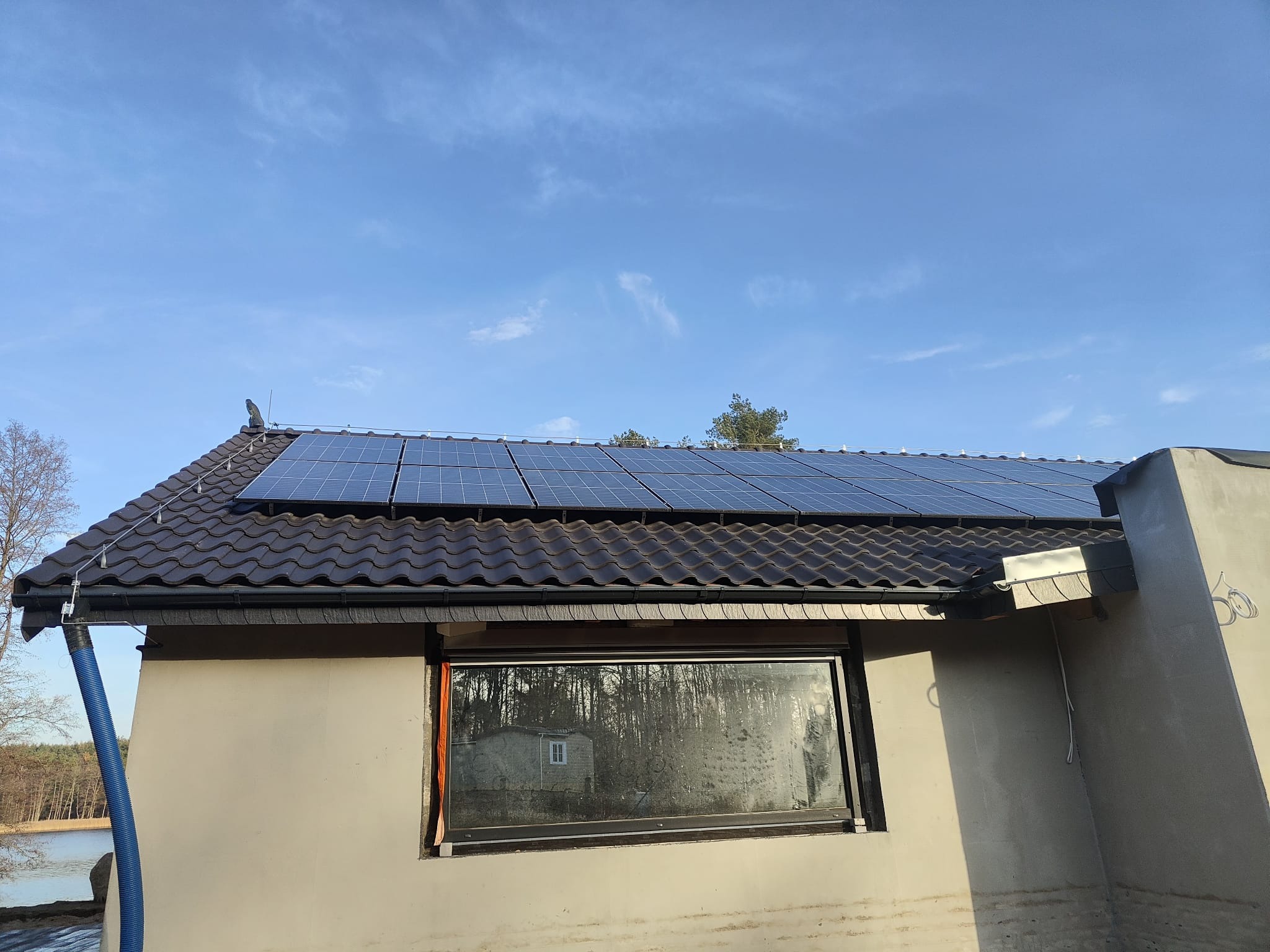 Panele słoneczne na dachu domu z ciemnej dachówki, widok z boku, okno z odbiciem drzew i niebieski wąż spustowy przy rynnie.