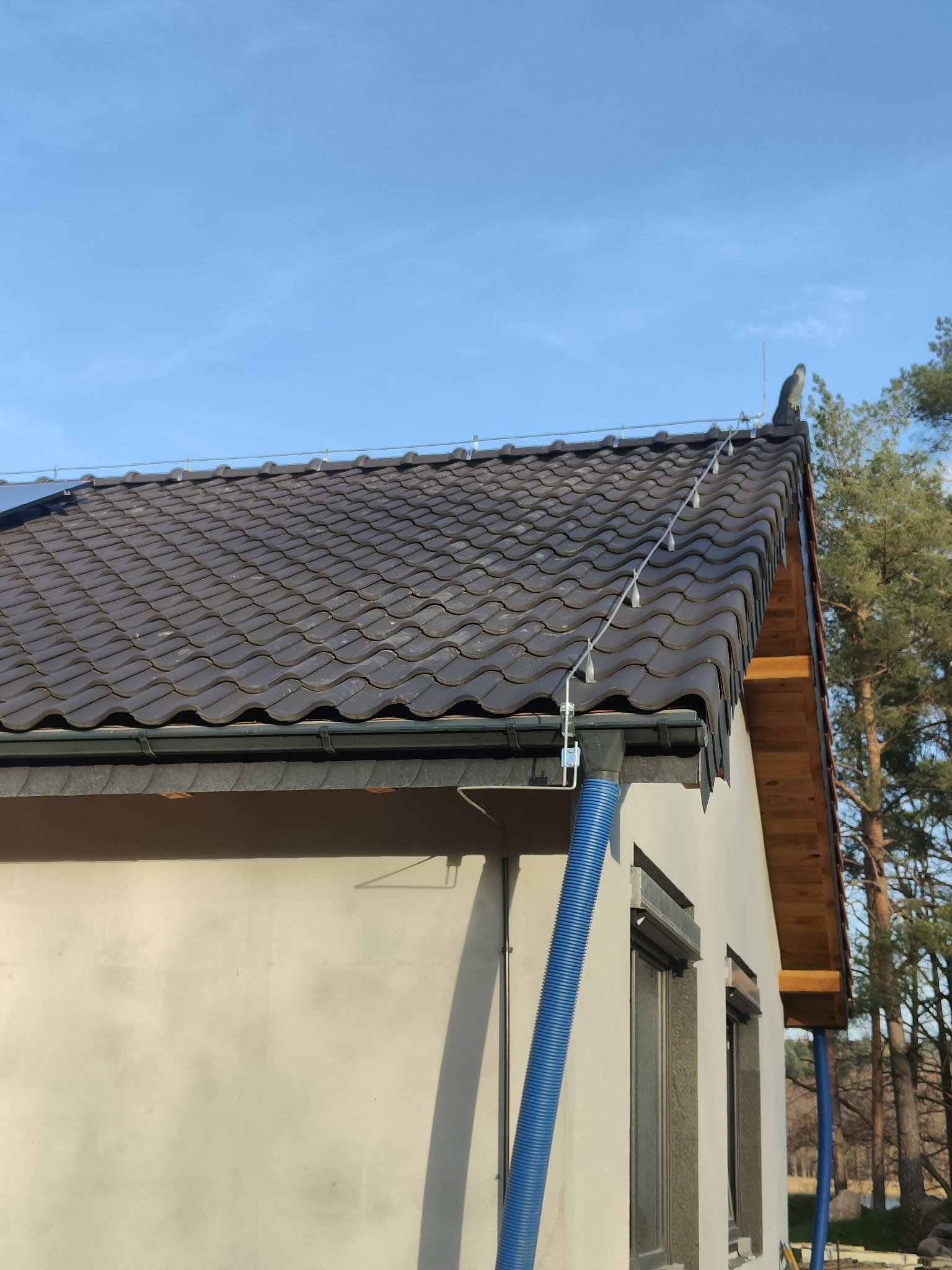 Nowy dom z brązowym dachem z dachówki ceramicznej i zainstalowaną instalacją odgromową z przewodami biegnącymi wzdłuż dachu, niebieski wąż wentylacyjny przy ścianie.