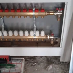 Rozdzielacz podłogowy z czerwonymi i białymi zaworami, zamontowany w obudowie, widoczne narzędzia i izolacja podłogowa na surowym podłożu.