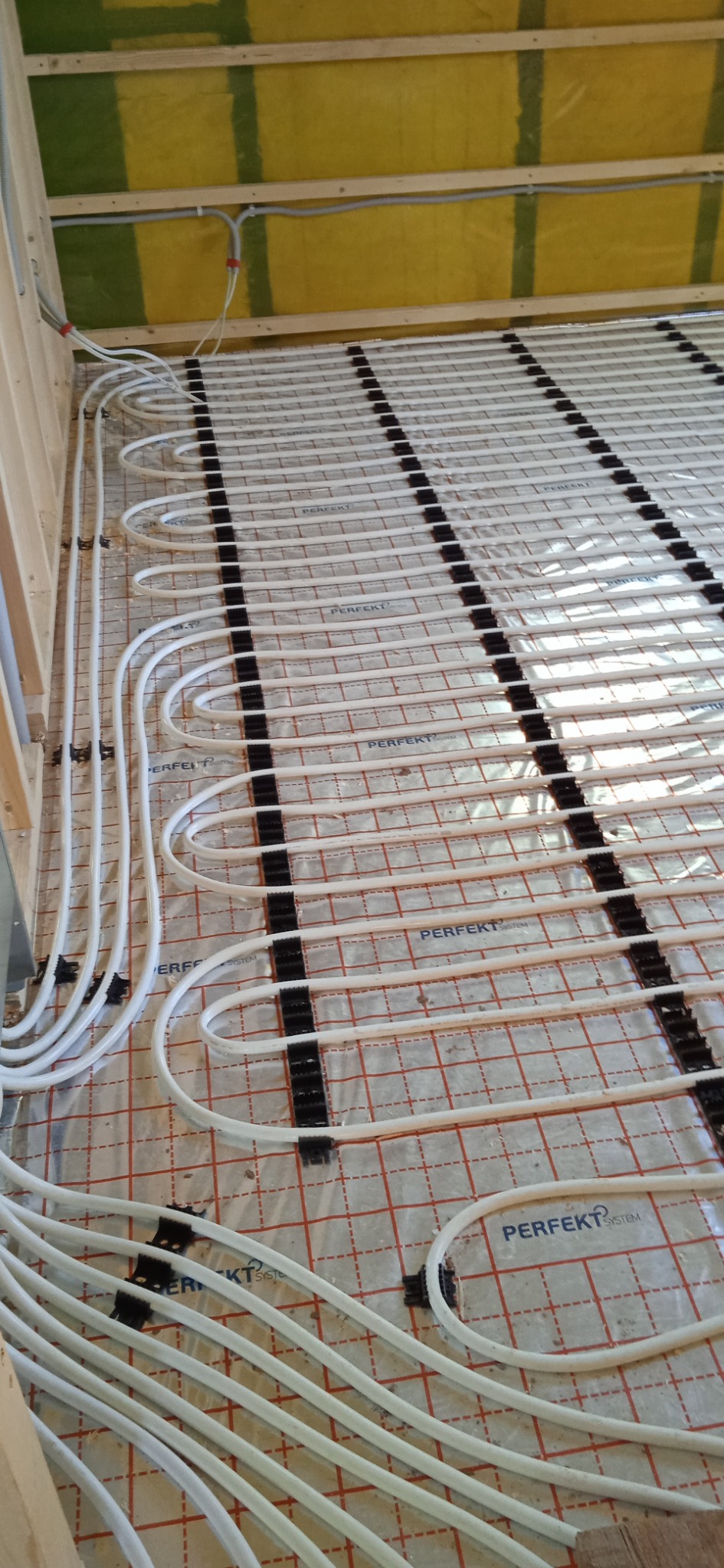 Białe rury ogrzewania podłogowego zamocowane czarnymi uchwytami na folii z nadrukiem 'PERFEKT SYSTEM', widoczne drewniane elementy konstrukcji budynku.