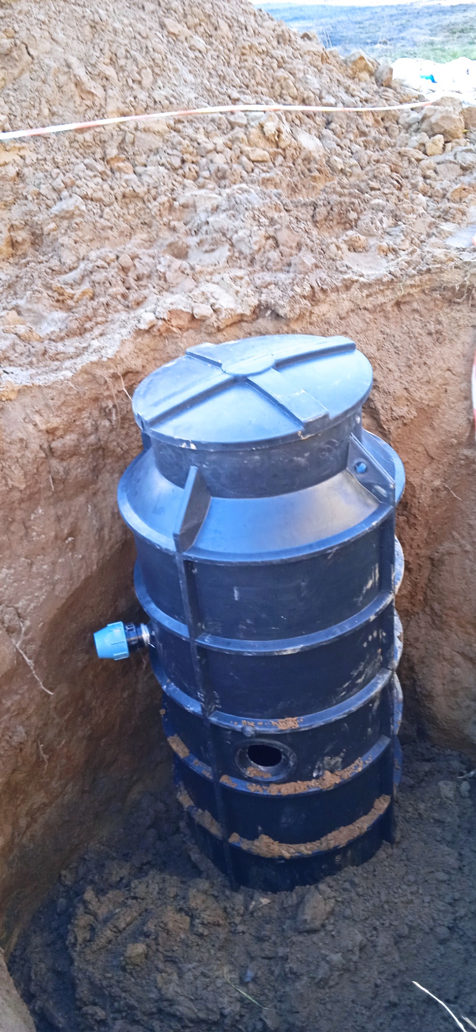 Czarny, plastikowy zbiornik bezodpływowy częściowo zakopany w wykopie ziemnym, widoczny niebieski króciec przyłączeniowy z boku.