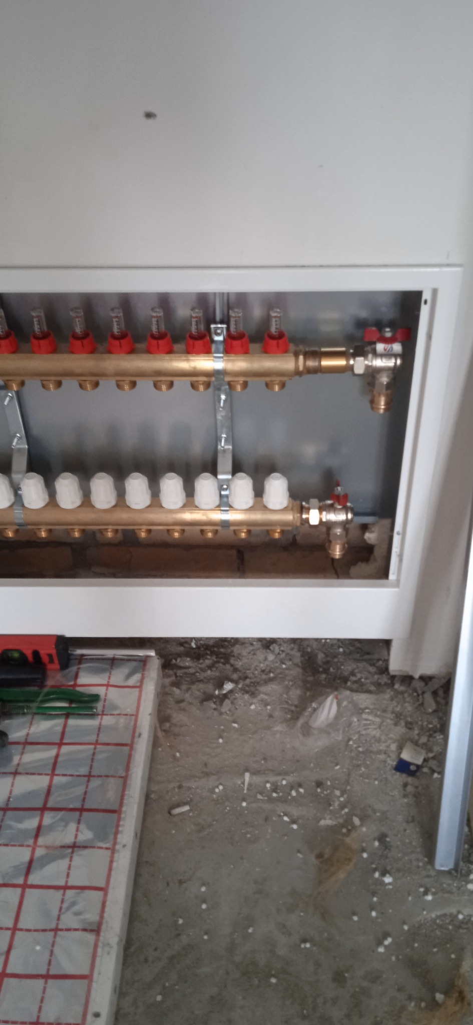 Rozdzielacz podłogowy z czerwonymi i białymi zaworami, zamontowany w obudowie, widoczne narzędzia i izolacja podłogowa na surowym podłożu.