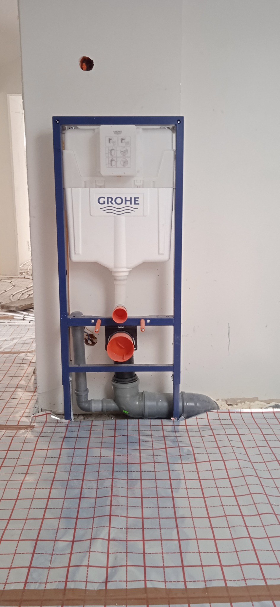 Stelaż podtynkowy Grohe zamontowany przy ścianie w surowym stanie, widoczne rury kanalizacyjne i ogrzewanie podłogowe w tle.