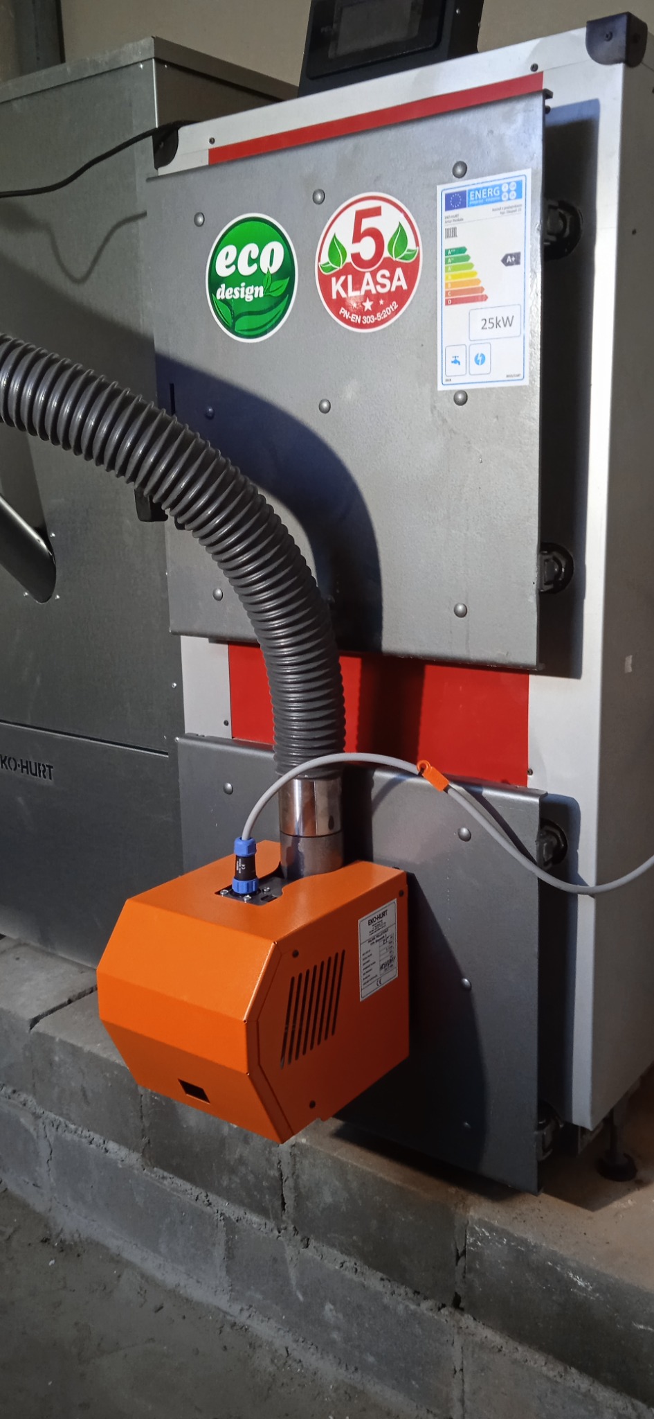 Szary piec CO z pomarańczowym podajnikiem, widoczne naklejki Eco Design i klasy 5, rura karbowana i etykieta energetyczna 25kW, zamontowany na betonowym fundamencie.