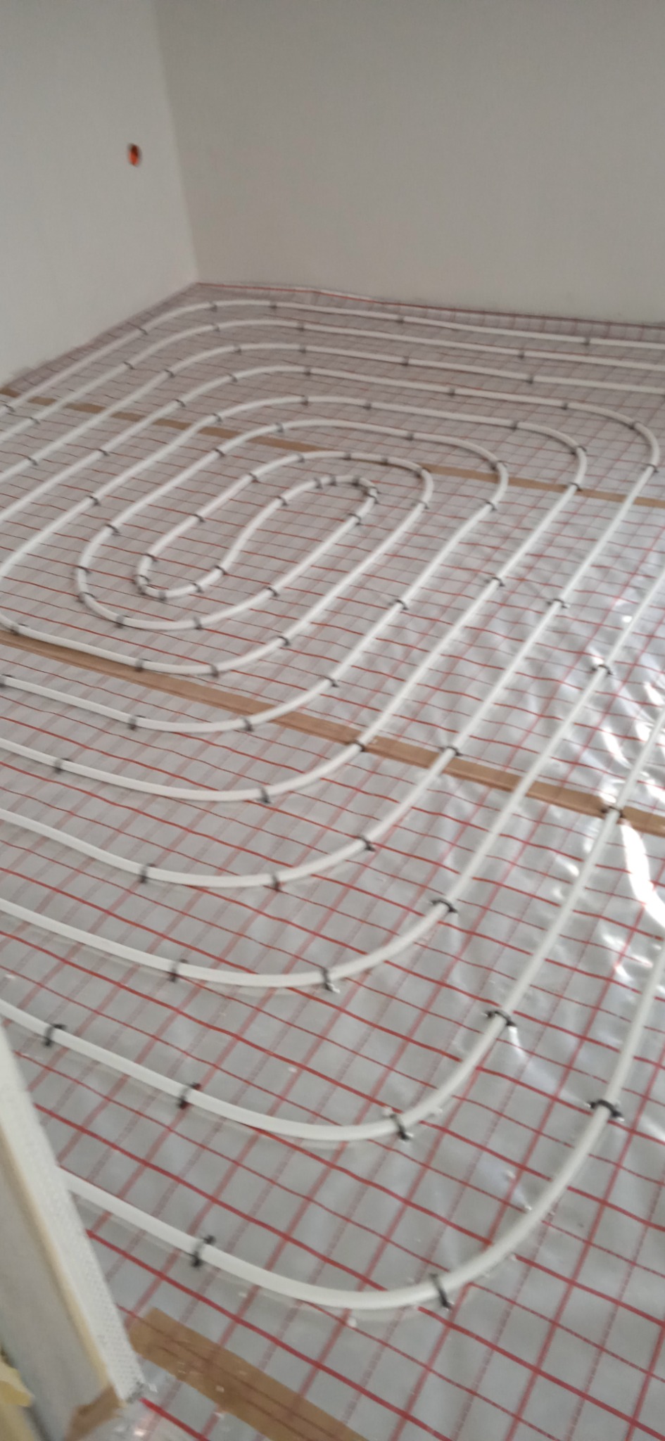 Białe rury ogrzewania podłogowego zamocowane na folii z czerwoną kratką, widok z góry na instalację w trakcie montażu, widoczne drewniane listwy mocujące.