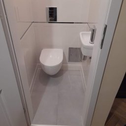 RENOVATION SYSTEMS Emil Oporski - Kompaktowa łazienka w bieli i szarościach z podwieszaną toaletą, małą umywalką narożną i szarą podłogą, widok zza otwartych drzwi na parkiet w jodełkę.