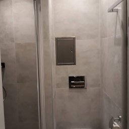 RENOVATION SYSTEMS Emil Oporski - Nowoczesna łazienka z szarą glazurą, kabiną prysznicową i podwieszaną toaletą. Widoczny metalowy właz rewizyjny i chromowany uchwyt na papier toaletowy.