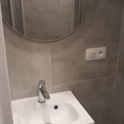 RENOVATION SYSTEMS Emil Oporski - Umywalka z szafką w szarej łazience, okrągłe lustro z widoczną kabiną prysznicową, gniazdko elektryczne na ścianie.