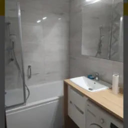 Nowoczesna łazienka z wanną z parawanem, umywalką nablatową na drewnianym blacie, pod kt&oacute;rym znajduje się pralka. Ściany wyłożone płytkami imitującymi naturalny kamień.