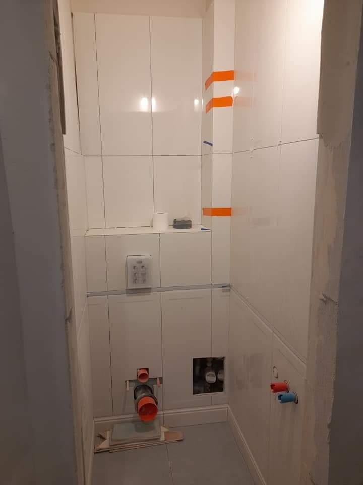 Wnętrze łazienki w trakcie prac wykończeniowych: białe płytki ułożone na ścianach, widoczne rury instalacyjne, taśmy malarskie zabezpieczające fugi i przyciski spłuczki w ścianie.