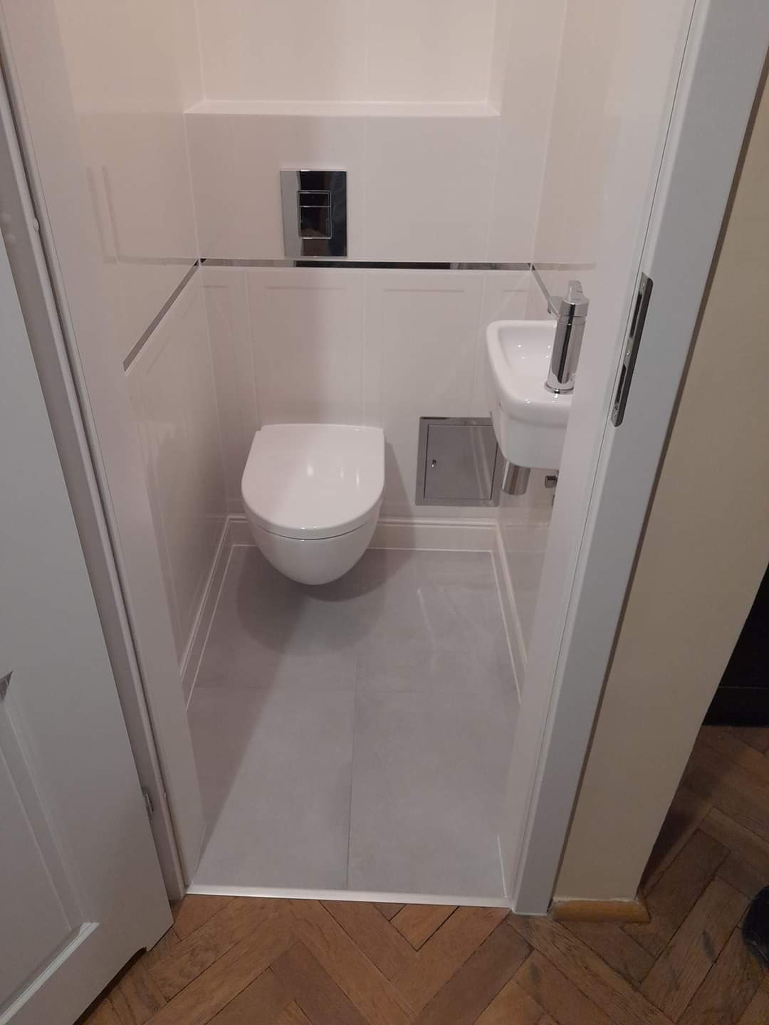 Kompaktowa łazienka w bieli i szarościach z podwieszaną toaletą, małą umywalką narożną i szarą podłogą, widok zza otwartych drzwi na parkiet w jodełkę.