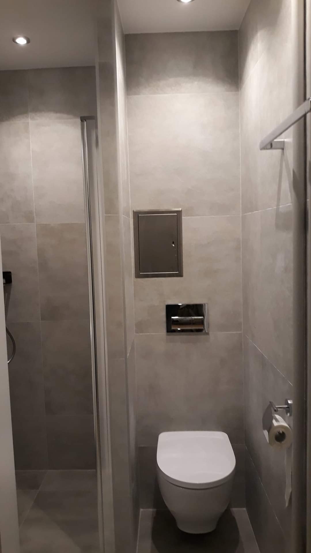 Nowoczesna łazienka z szarą glazurą, kabiną prysznicową i podwieszaną toaletą. Widoczny metalowy właz rewizyjny i chromowany uchwyt na papier toaletowy.