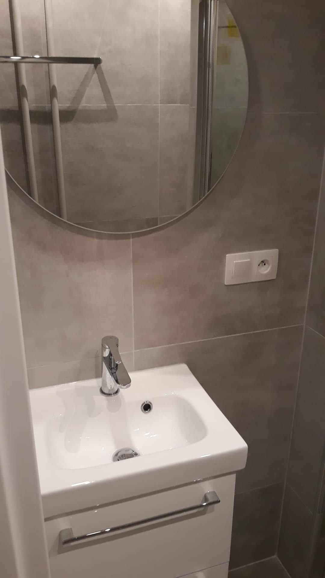 Umywalka z szafką w szarej łazience, okrągłe lustro z widoczną kabiną prysznicową, gniazdko elektryczne na ścianie.