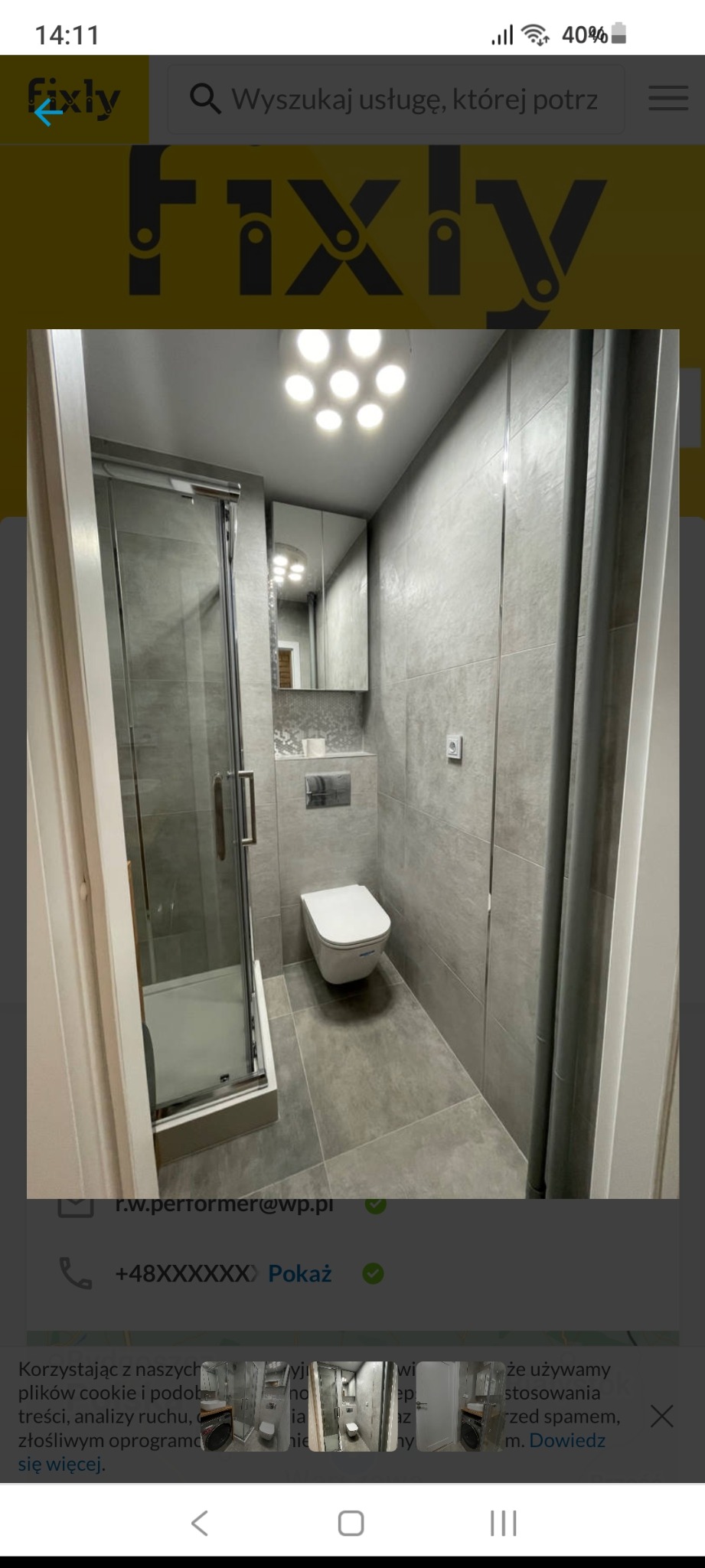 Wąska łazienka po odnowieniu, z kabiną prysznicową ze szklanymi drzwiami, podwieszaną toaletą i szafką z lustrem, wykończona szarymi płytkami, oświetlona punktowymi lampami sufitowymi.