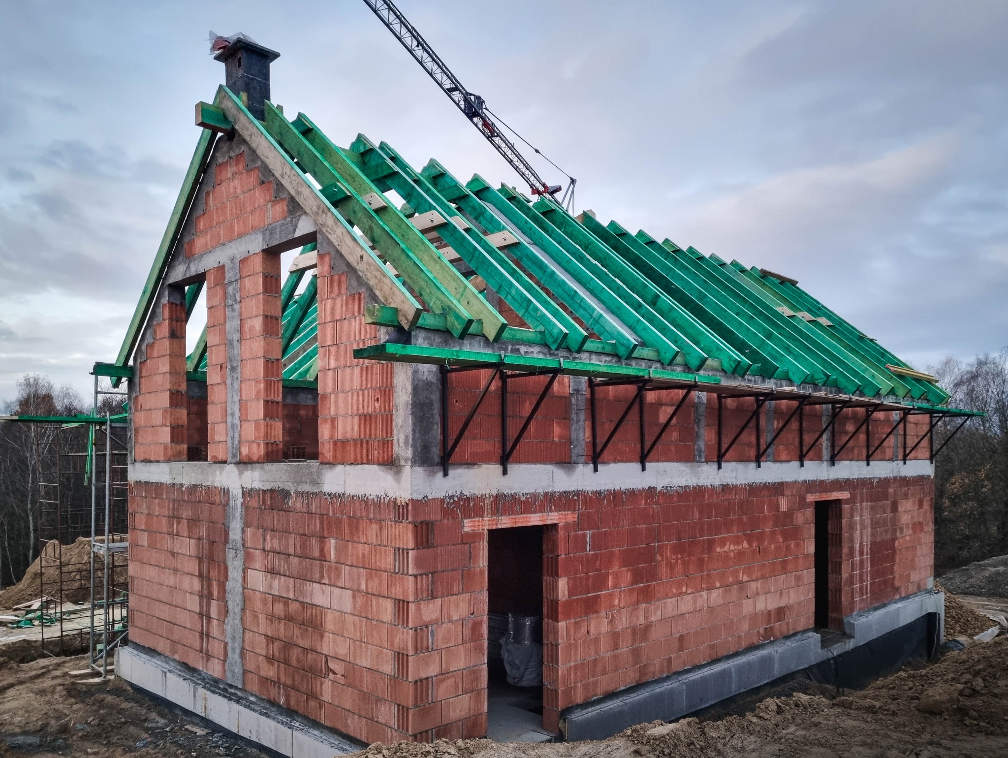 Surowa bryła domu w stanie deweloperskim, ściany z czerwonej cegły, zielona konstrukcja dachu, widoczny dźwig budowlany w tle.