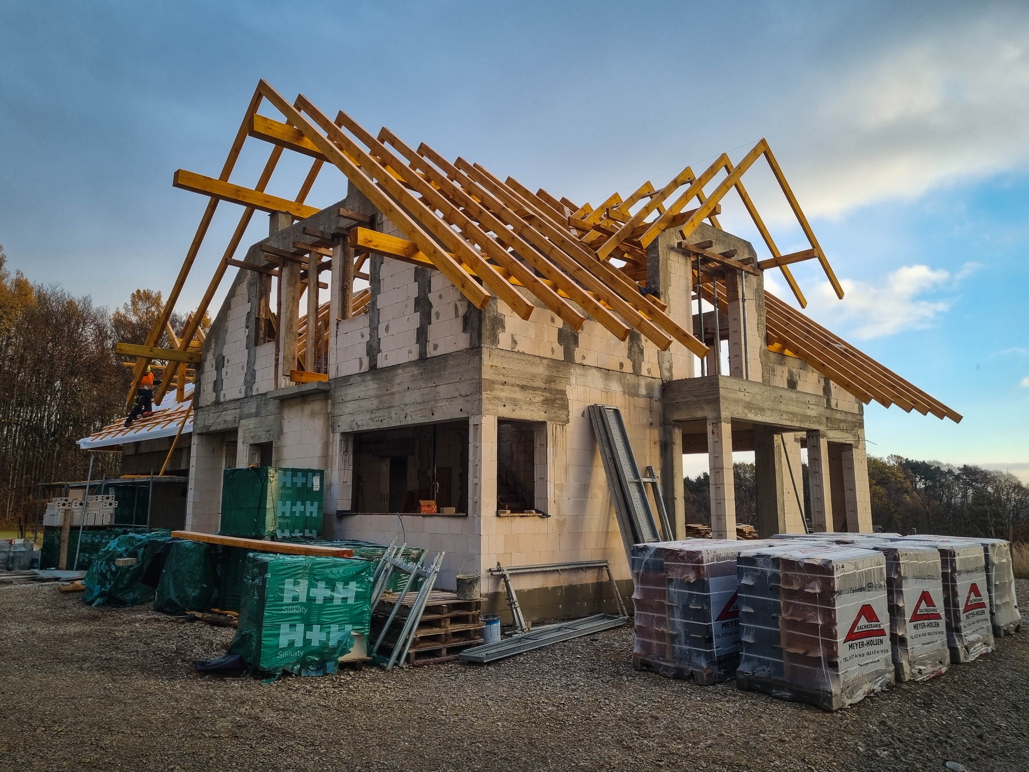 Budowa domu jednorodzinnego w Krakowie w stanie surowym otwartym, widoczne ściany z bloczków silikatowych, drewniana konstrukcja dachu, materiały budowlane na paletach i pracownik w kasku na dachu.