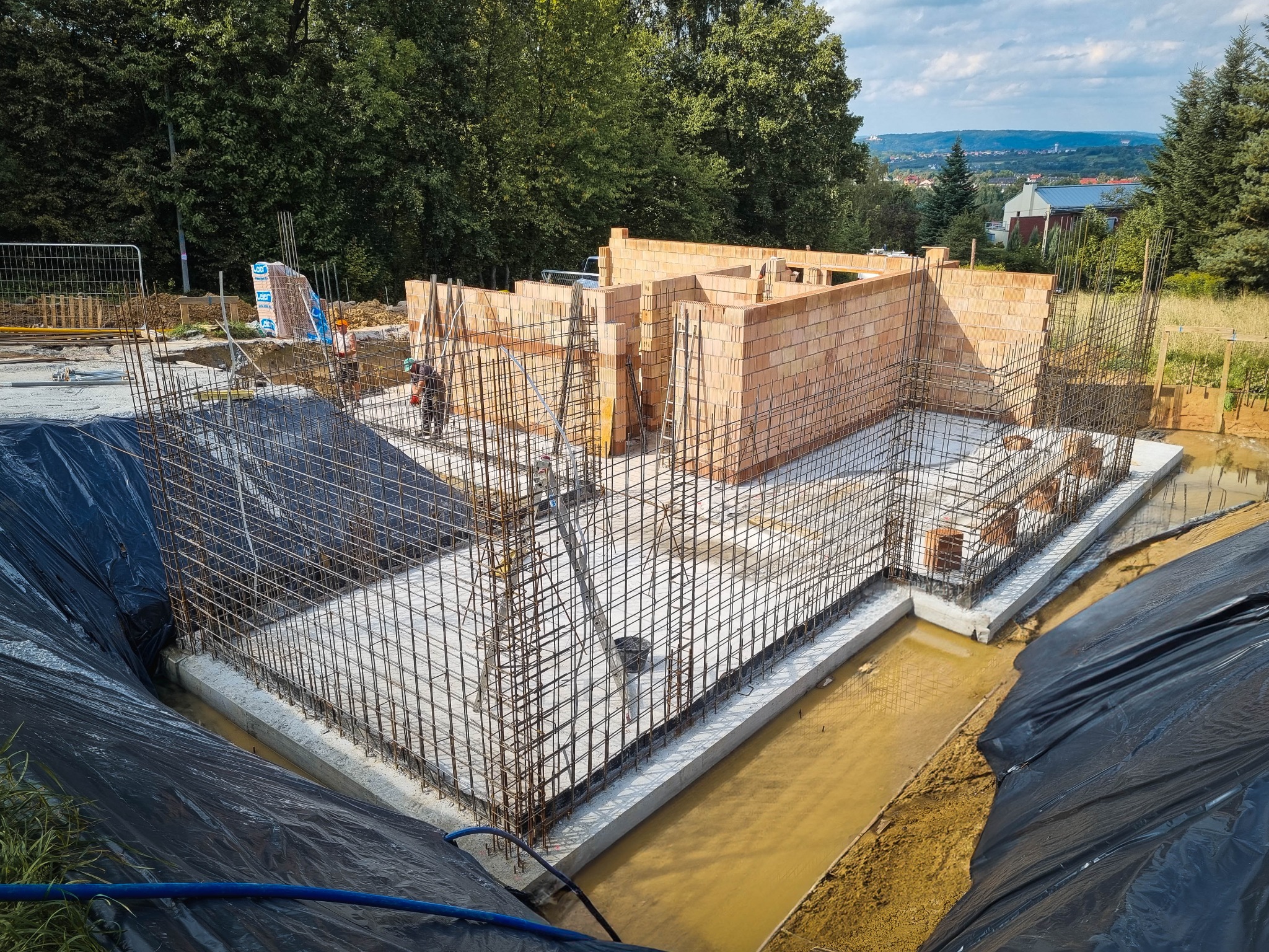 Budowa domu jednorodzinnego w stanie surowym otwartym: wylany fundament, zbrojenie, pierwsze warstwy muru z bloczków, widok na okolicę w tle.