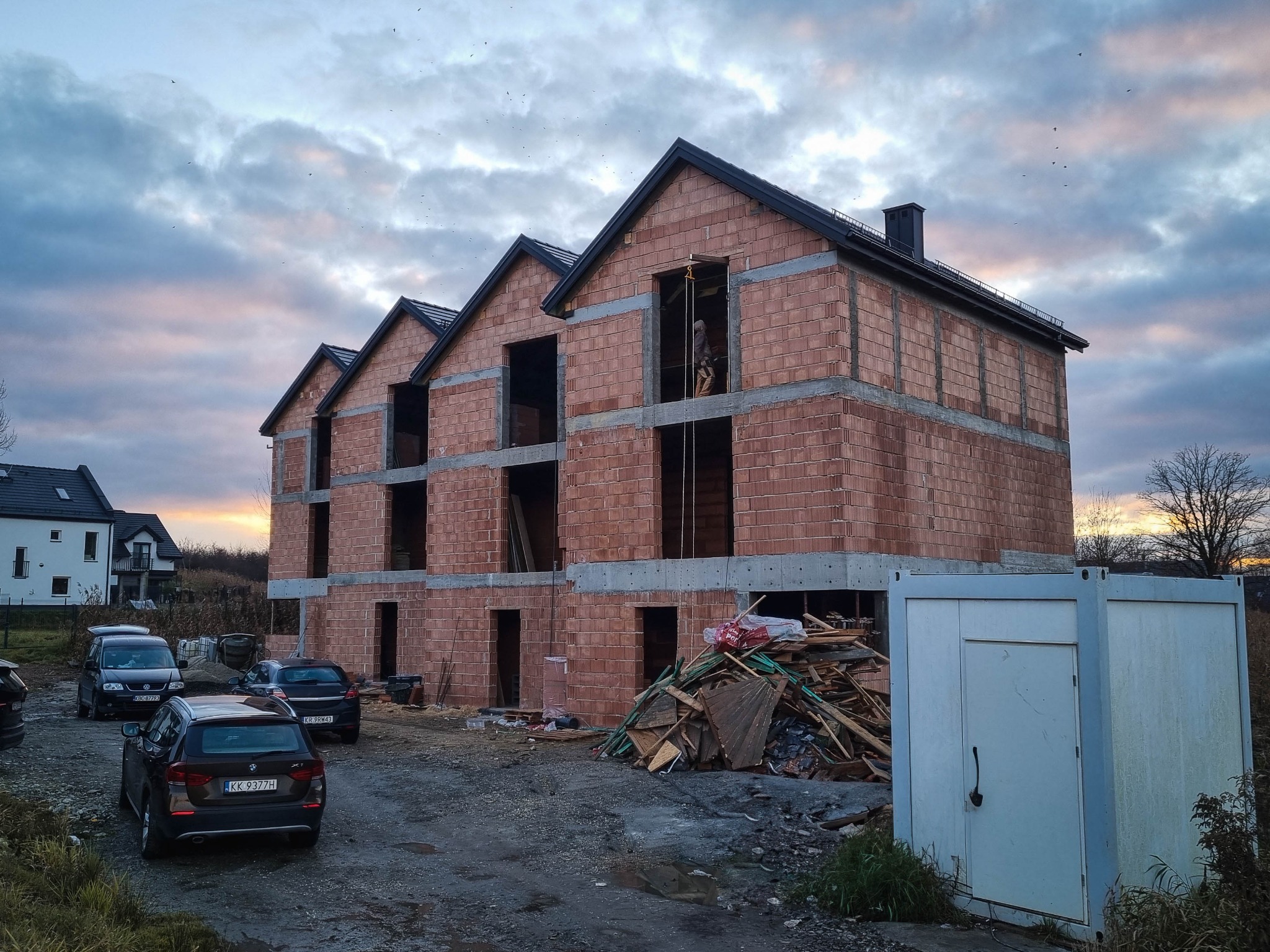 Surowy stan trzypiętrowego budynku mieszkalnego z czerwonej cegły, z widocznym pracownikiem w oknie na drugim piętrze, na tle pochmurnego nieba o zmierzchu.