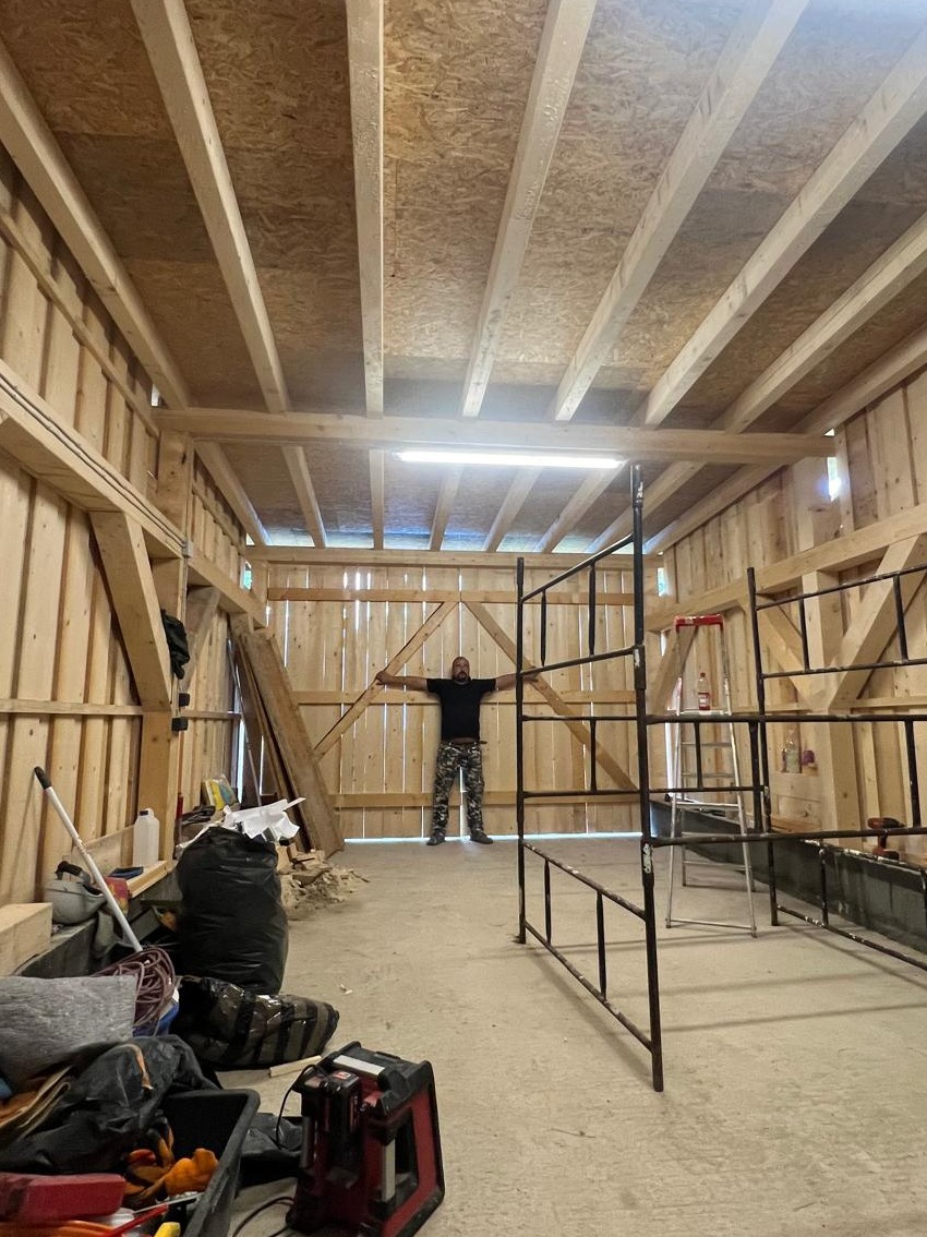 Wnętrze drewnianej konstrukcji szkieletowej w trakcie budowy, z widocznymi belkami stropowymi, deskami ściennymi, mężczyzną w tle oraz narzędziami i workami z materiałami budowlanymi na podłodze.