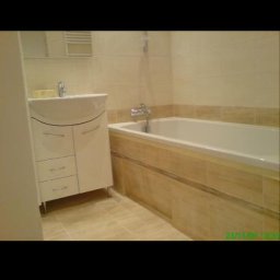 remontik - Jasna łazienka z wanną obudowaną płytkami imitującymi drewno, białą szafką umywalkową i beżowymi płytkami na ścianach.