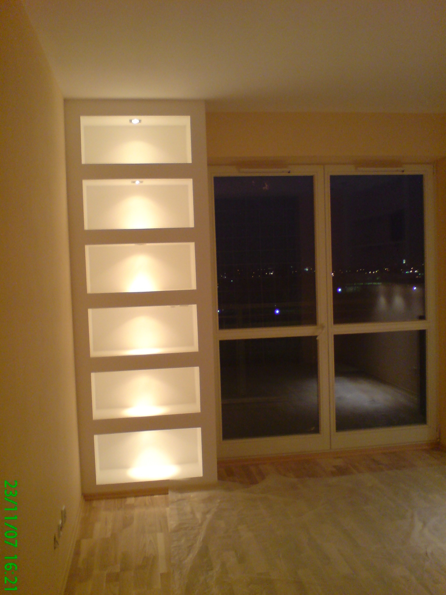 Wnęka ścienna z sześcioma podświetlanymi półkami obok okna balkonowego z widokiem na nocne miasto, parkiet częściowo zakryty folią ochronną.