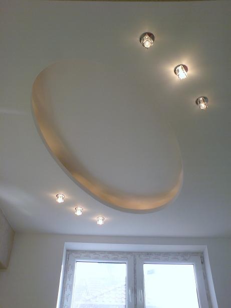 Biały sufit podwieszany z owalnym wgłębieniem oświetlonym ciepłym światłem, uzupełniony punktowymi lampami halogenowymi, widok z dołu na tle okna.