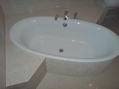 Owalna, biała wanna wolnostojąca z chromowaną armaturą, obudowana mozaiką w odcieniach szarości, na tle płytek podłogowych w kolorze beżowym.