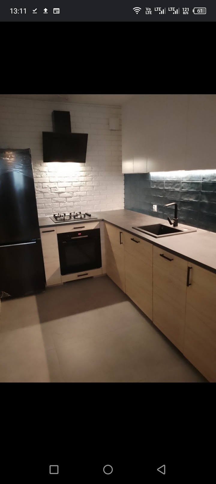 Nowoczesna kuchnia z białymi cegłami na ścianie, jasnymi drewnianymi szafkami z czarnymi uchwytami, czarnym zlewem i czarną armaturą oraz czarnym okapem i lodówką.