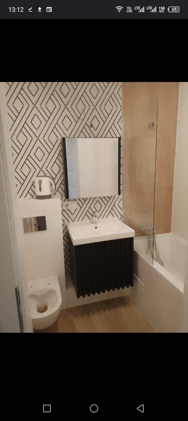 Nowoczesna łazienka z geometryczną tapetą, czarną szafką pod umywalką, wiszącą toaletą i parawanem nawannowym ze szkła hartowanego.