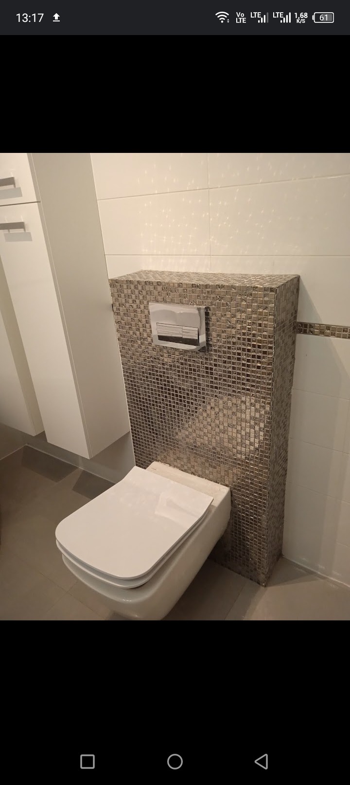 Podwieszana toaleta z obudową ze srebrnej mozaiki, chromowany przycisk spłukiwania, białe płytki na ścianie i szare na podłodze, minimalistyczna szafka w tle.