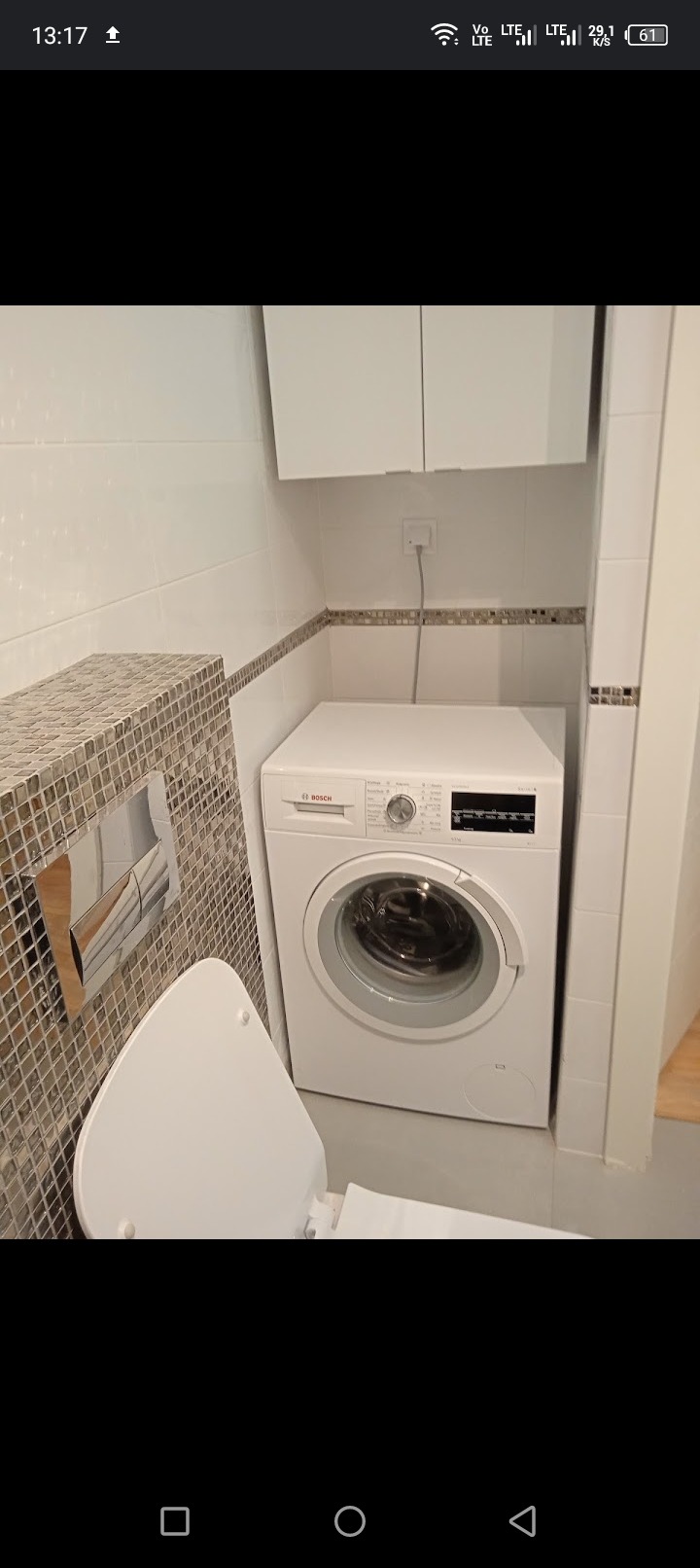 Kompaktowa łazienka z białą pralką Bosch w zabudowie, mozaika na ścianie i podwieszana szafka nad pralką, widoczny otwarty sedes.