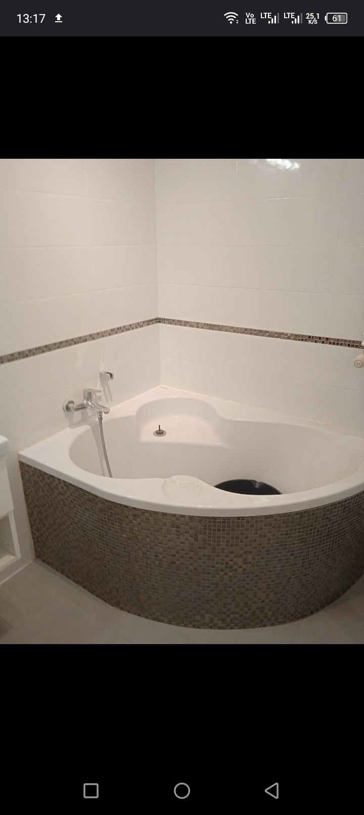 Narożna wanna obudowana mozaiką w łazience z białymi płytkami i dekoracyjnym paskiem mozaiki wzdłuż ściany, widoczna bateria i odpływ.