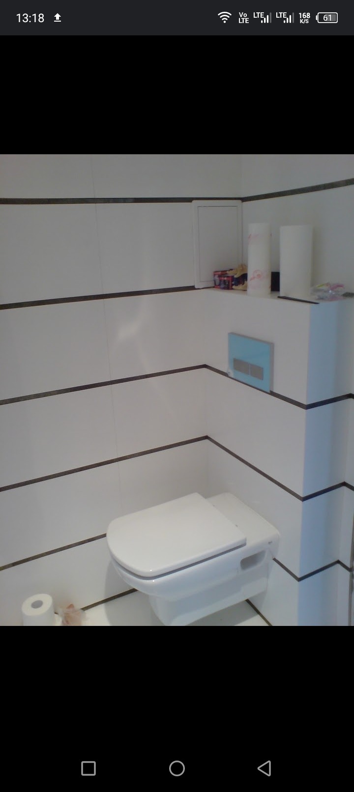 Nowoczesna toaleta podwieszana z niebieskim panelem spłukującym, białe płytki z poziomymi, ciemnymi fugami, dodatki na półce nad toaletą.