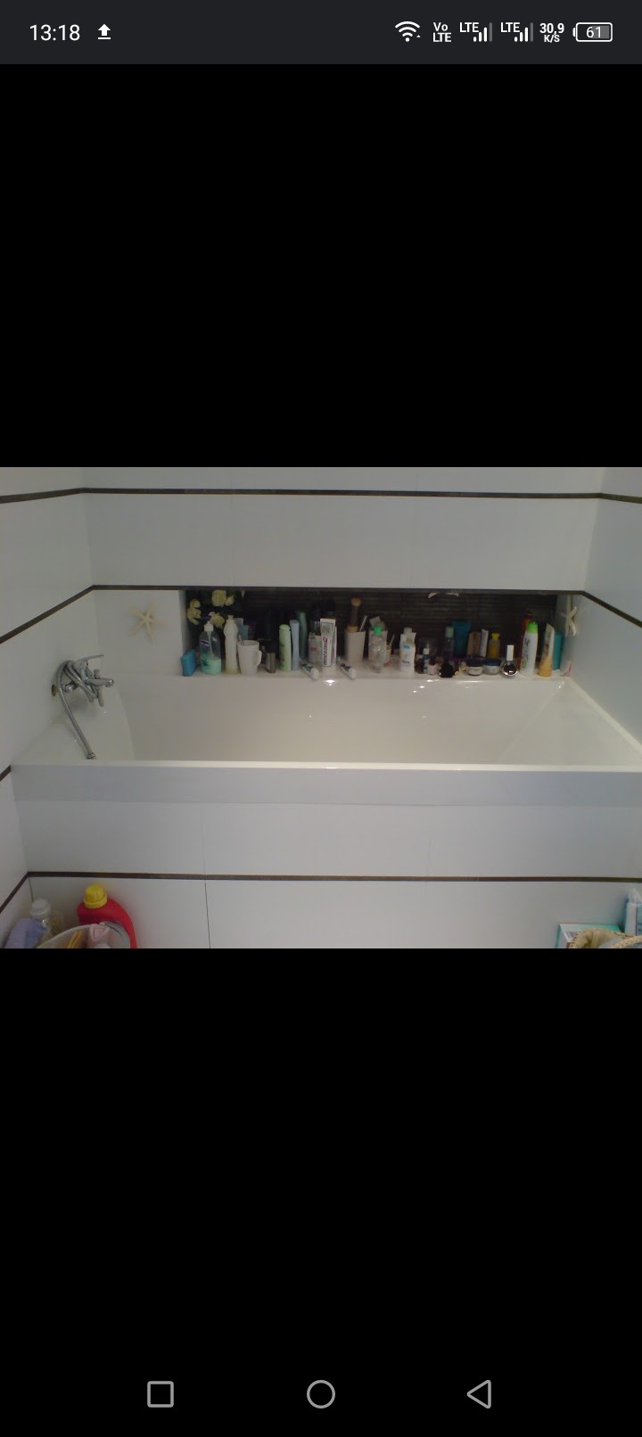 Biała wanna z baterią i półką w zabudowie, na której stoją liczne kosmetyki i akcesoria łazienkowe, a na ścianie wisi dekoracyjna gwiazda morska.