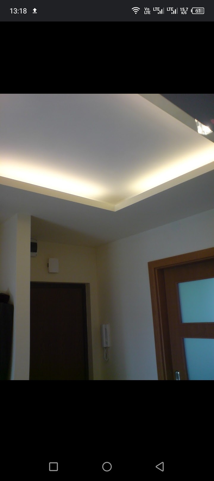 Nowoczesny sufit podwieszany z oświetleniem LED w narożniku, widok na fragment pokoju z drzwiami i domofonem na ścianie.