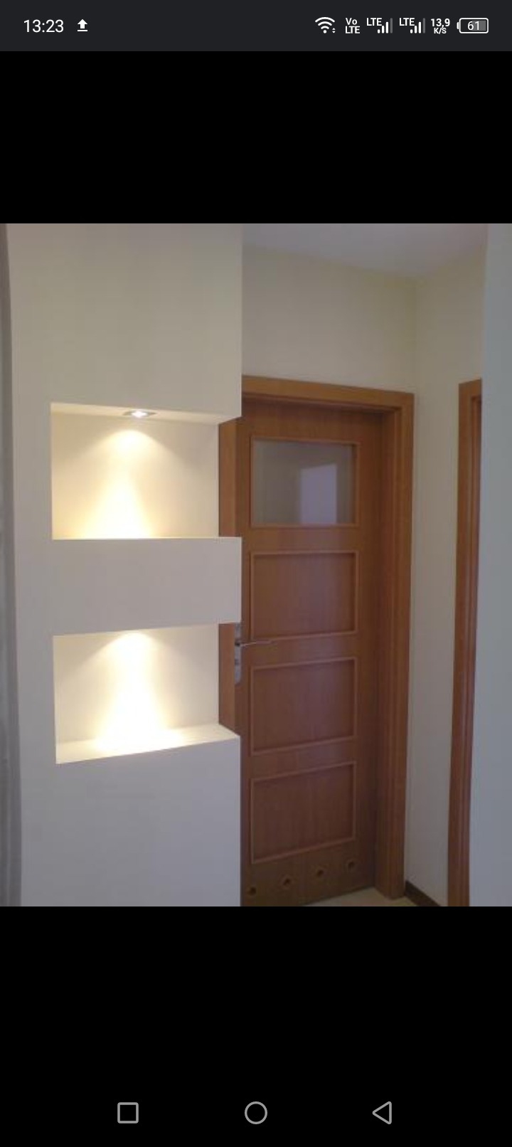 Nowoczesna wnęka ścienna z dwoma podświetlonymi półkami, obok drewniane drzwi wewnętrzne z szybą, kremowe ściany.